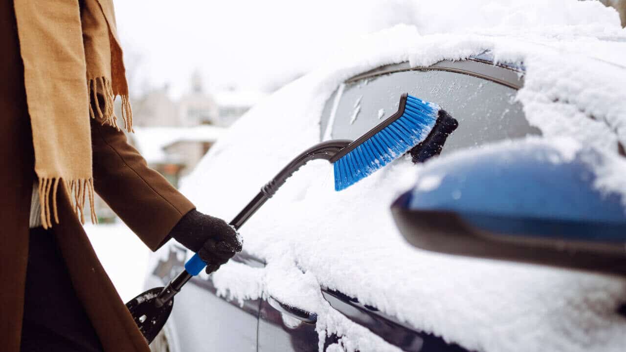 Person in a coat and scarf clears snow from his car during a winter snowfall. Seasonality concept, weather, people, transport.