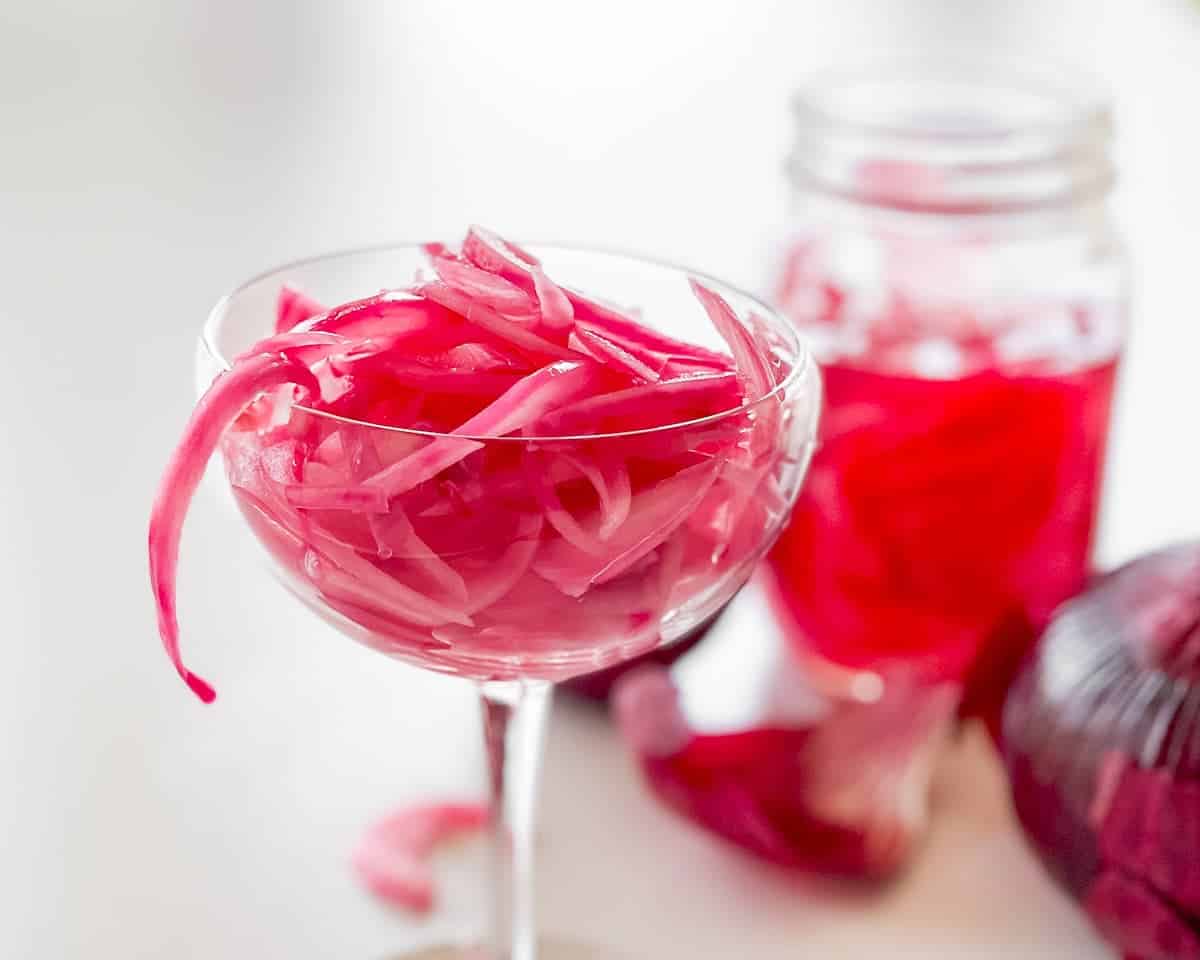 Easy pickled red onions i a coupe glass with a jar full of them and uncut red onions in the background.