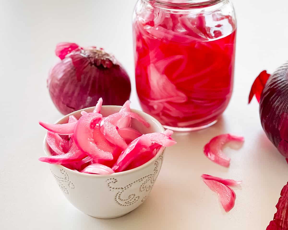 Red onions that have been pickled in a white bowl. A jar full of pickled onions and whole red onions shown nearby. All on a white table seen from a side angle.